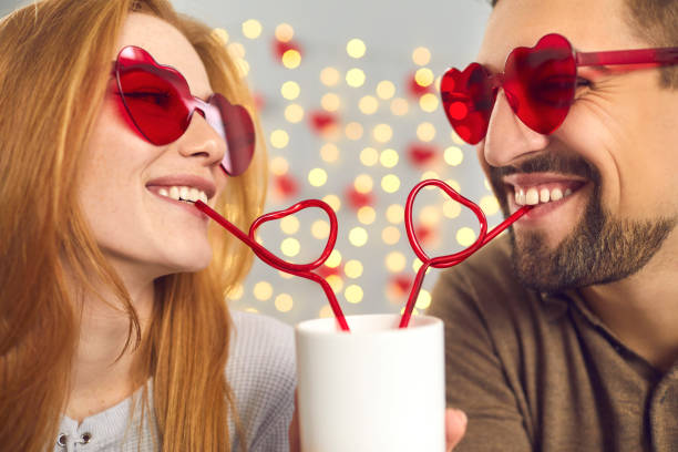 Close-up portrait happy young man and woman in love in pink sunglasses sipping drink from one cup through heart-shaped straws, enjoying cute funny couple moment on a fun date on Saint Valentine's Day